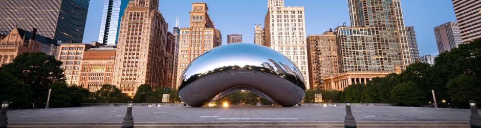 Die Cloud-Gate-Skulptur, auch bekannt als The Bean, im Millennium Park in Chicago spiegelt bei Sonnenaufgang die nahe gelegenen Bäume und Wolkenkratzer der Stadt vor einem klaren Himmel im Hintergrund wider.
