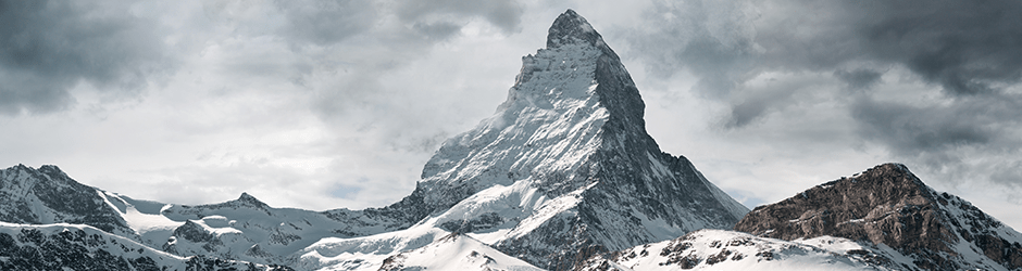 Schneebedeckter Berggipfel unter bewölktem Himmel, mit scharfen Graten und felsigen Hängen, die teilweise mit Schnee bedeckt sind. Kleinere Berge und Bergrücken erscheinen im Vordergrund.