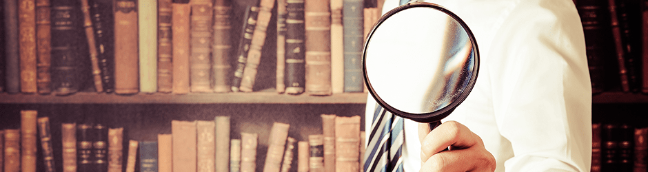 Eine Person in Hemd und Krawatte hält ein Vergrößerungsglas vor ein mit alten Büchern gefülltes Bücherregal, das ihr Gesicht teilweise verdeckt.