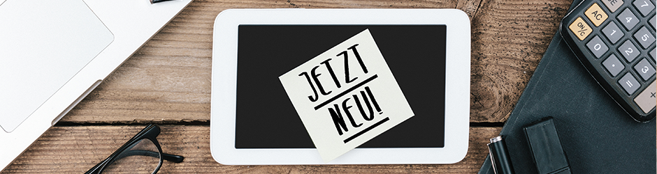 Ein Klebezettel mit dem Text "JETZT NEU!" befindet sich auf einem Tablet-Bildschirm, umgeben von einem Laptop, einem Taschenrechner, einer Brille, einem Stift und einem Ordner auf einem Holztisch.