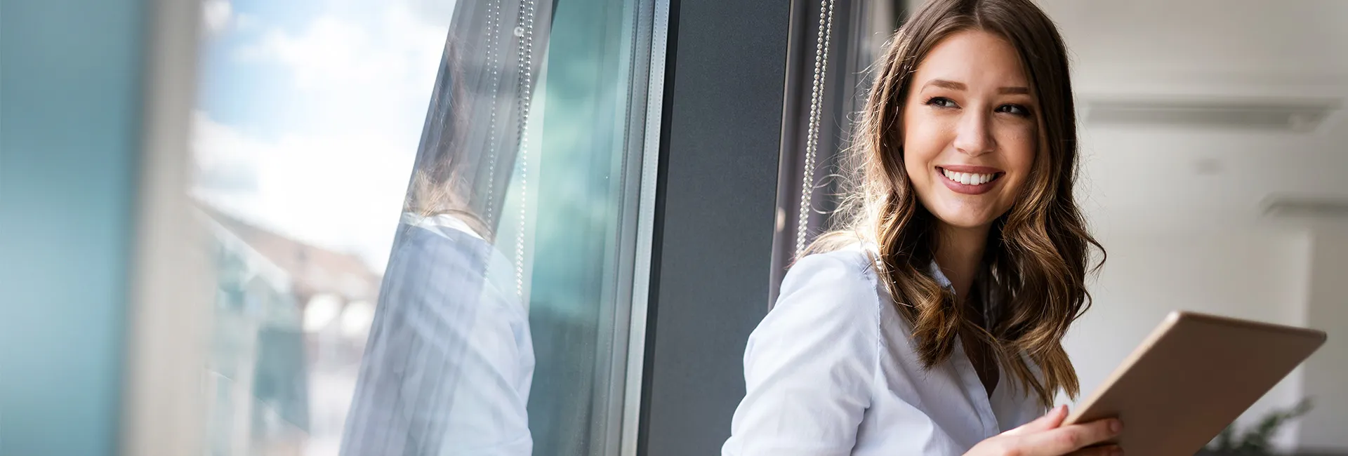 Eine junge Frau mit langen braunen Haaren lächelt und hält ein Tablet. Sie sitzt vor einem großen Fenster.